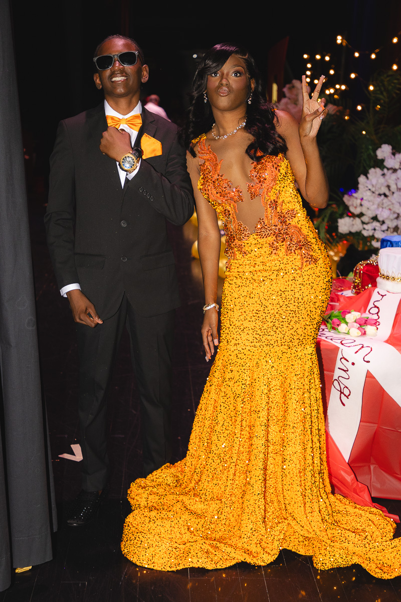 Man in a black suit with an orange bow tie and sunglasses poses with a woman in a bright yellow sequined gown at a formal event.