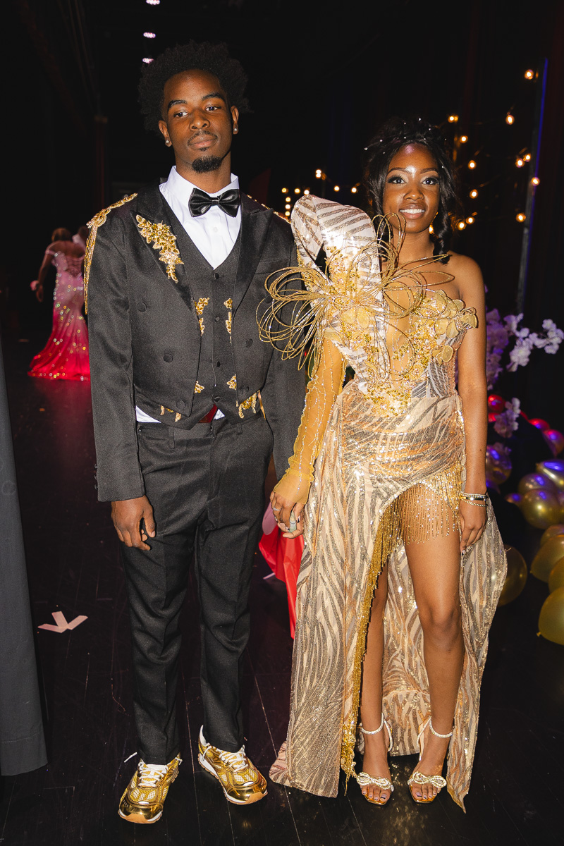 Couple in formal attire posing backstage at a gala; man in black suit with gold accents and bow tie, woman in gold embellished gown holding his hand.