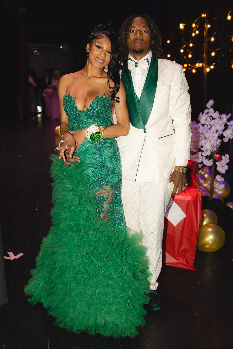Couple in formal wear posing together: woman in a emerald green ball gown with ruffled skirt and man in a white suit with green accents and bow tie.