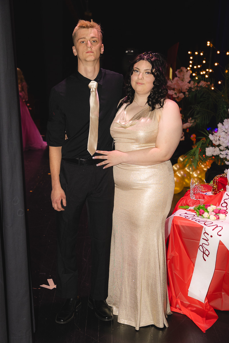Young man in a black shirt with a beige tie stands beside a woman in a champagne-colored gown at a formal event, posing for a photo.