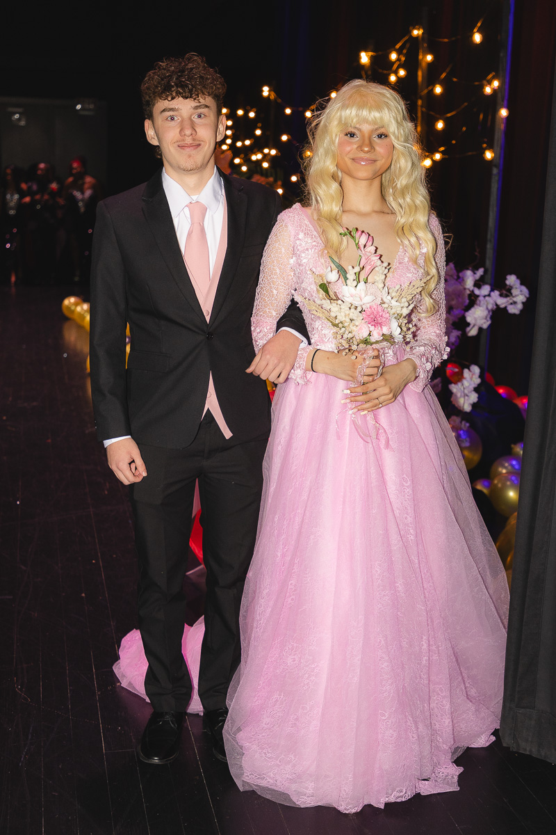 Pair of teens in formal wear at prom: boy in a black suit with a pink tie and girl in a pink ball gown holding a bouquet, standing together with lights in the background.