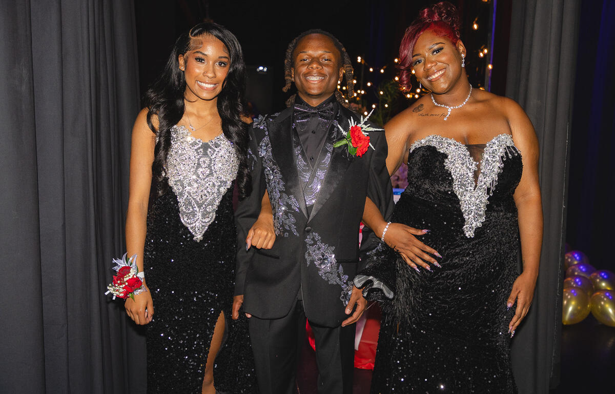 Three formally dressed friends posing together at an event, smiling at the camera in black and silver gowns and a suit with boutonniere.