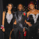 Three formally dressed friends posing together at an event, smiling at the camera in black and silver gowns and a suit with boutonniere.