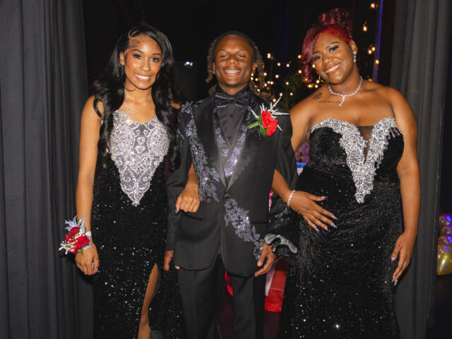Three formally dressed friends posing together at an event, smiling at the camera in black and silver gowns and a suit with boutonniere.
