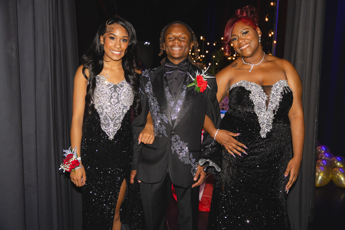 Three formally dressed friends posing together at an event, smiling at the camera in black and silver gowns and a suit with boutonniere.