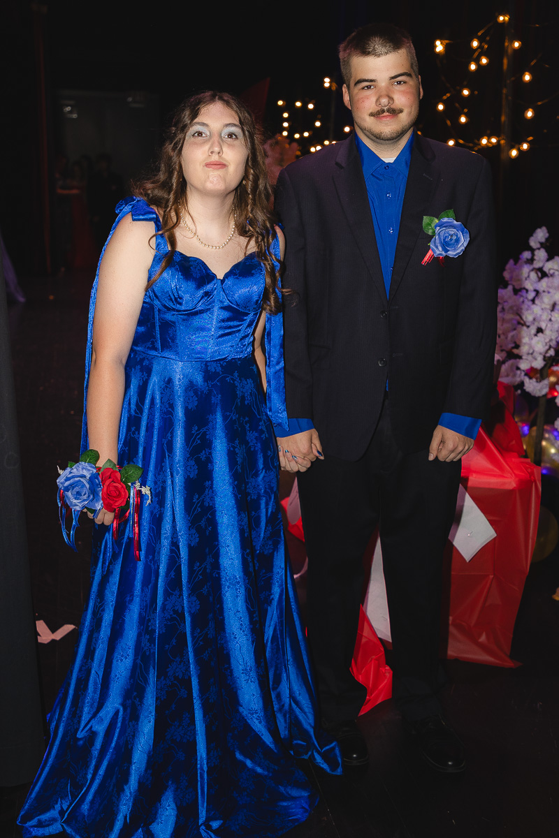 Couple at a formal event holding hands; woman in a royal blue ball gown with a corsage, man in a dark suit with a blue shirt and boutonniere.