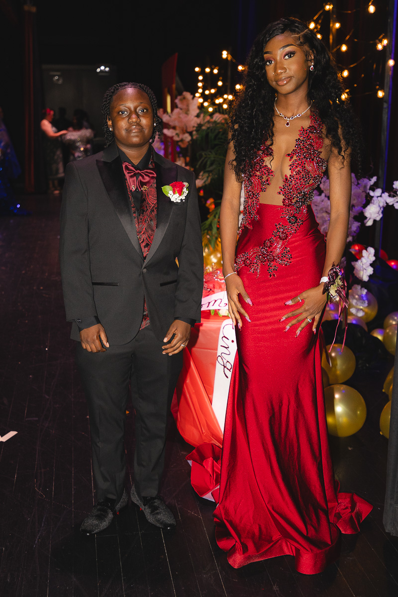 Two teens in formal wear posing on a stage: a boy in a black suit with burgundy vest and bow tie, corsage on lapel; a girl in a red beaded gown posing beside him.