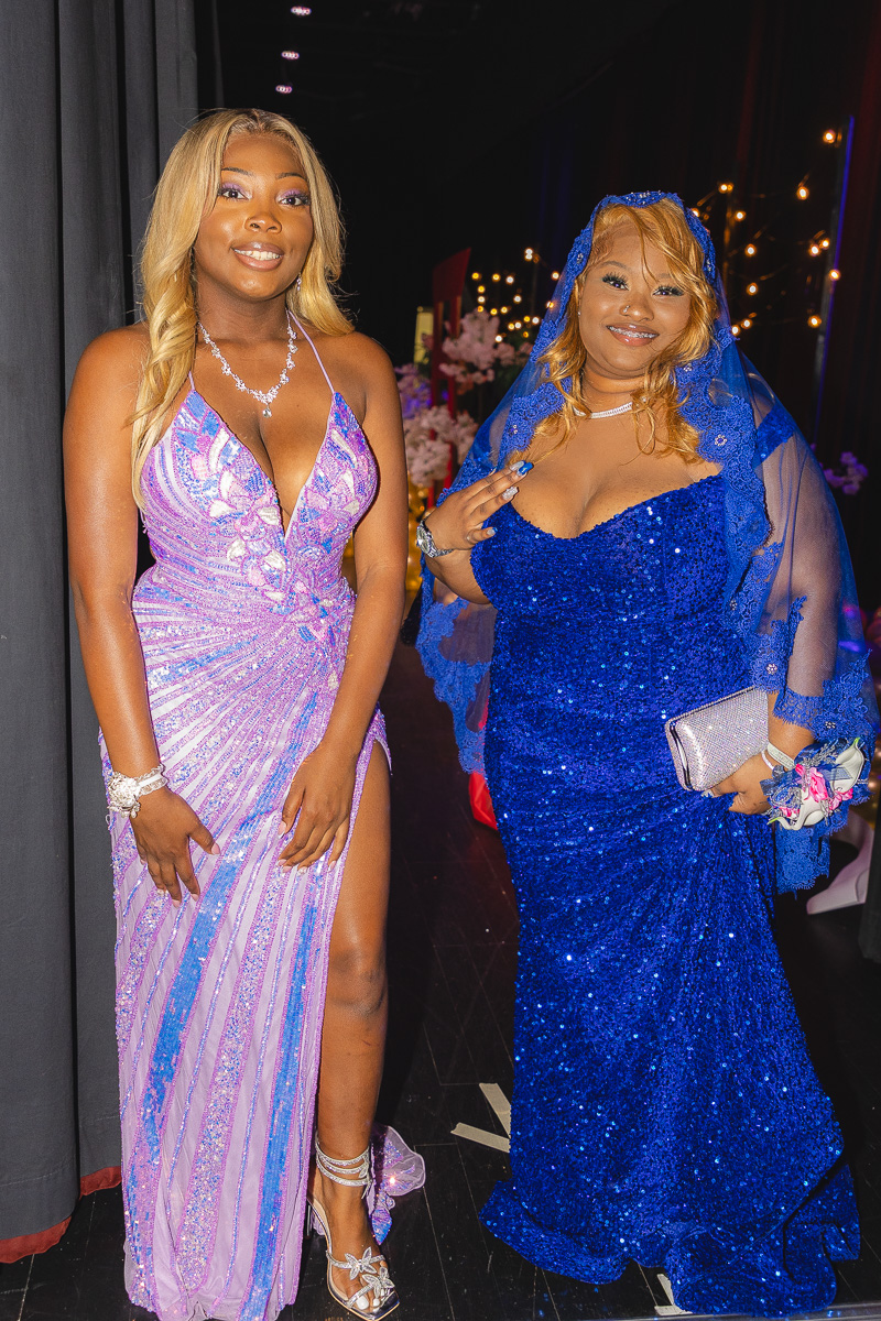 Two women in glamorous gowns pose for a photo at an event; one wears a lavender sequin dress, the other a blue gown with a matching veil.