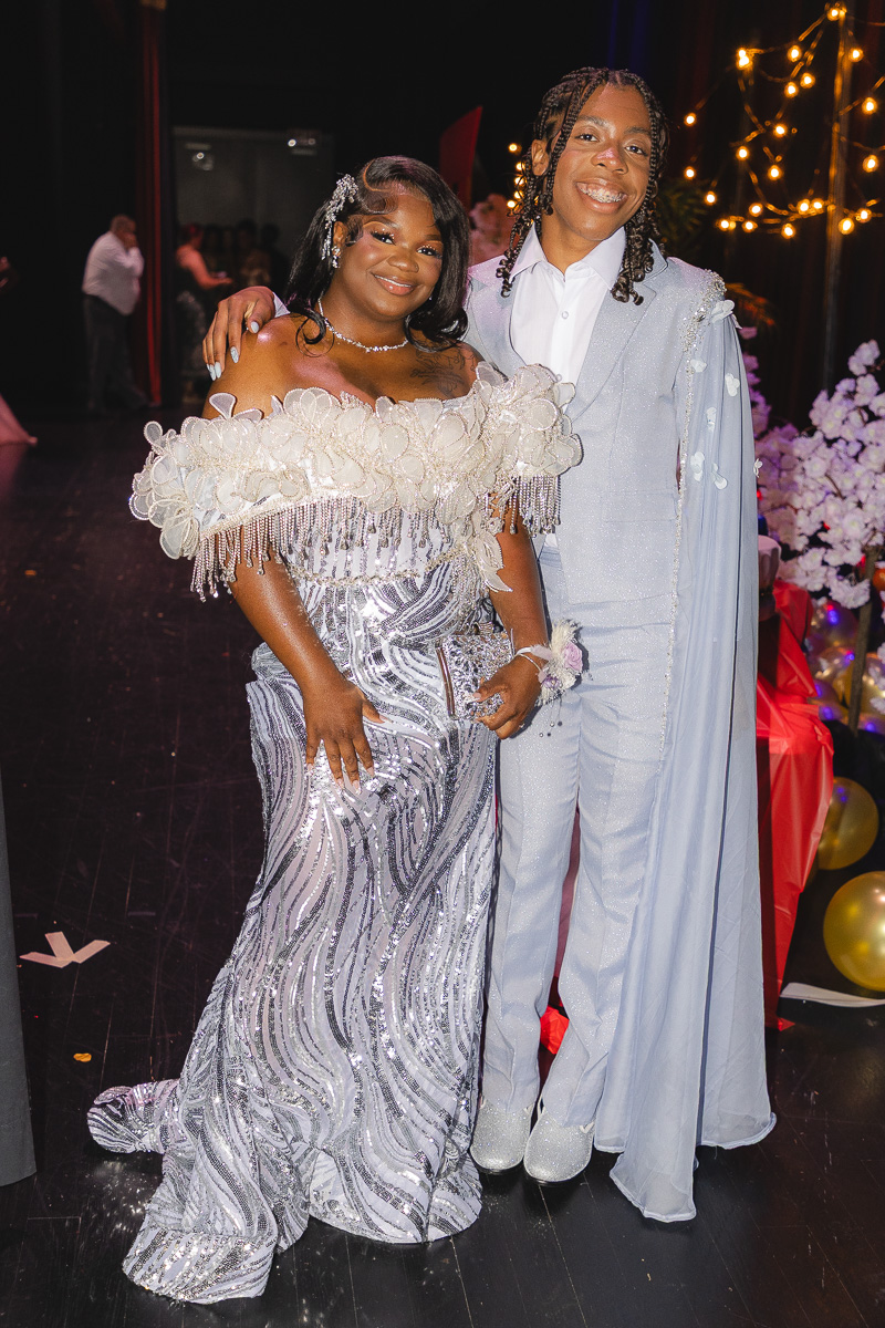 Two young people in formal attire pose together at a party, the woman wearing a silver off‑the‑shoulder sequined gown and the man in a pale blue suit with a cape.