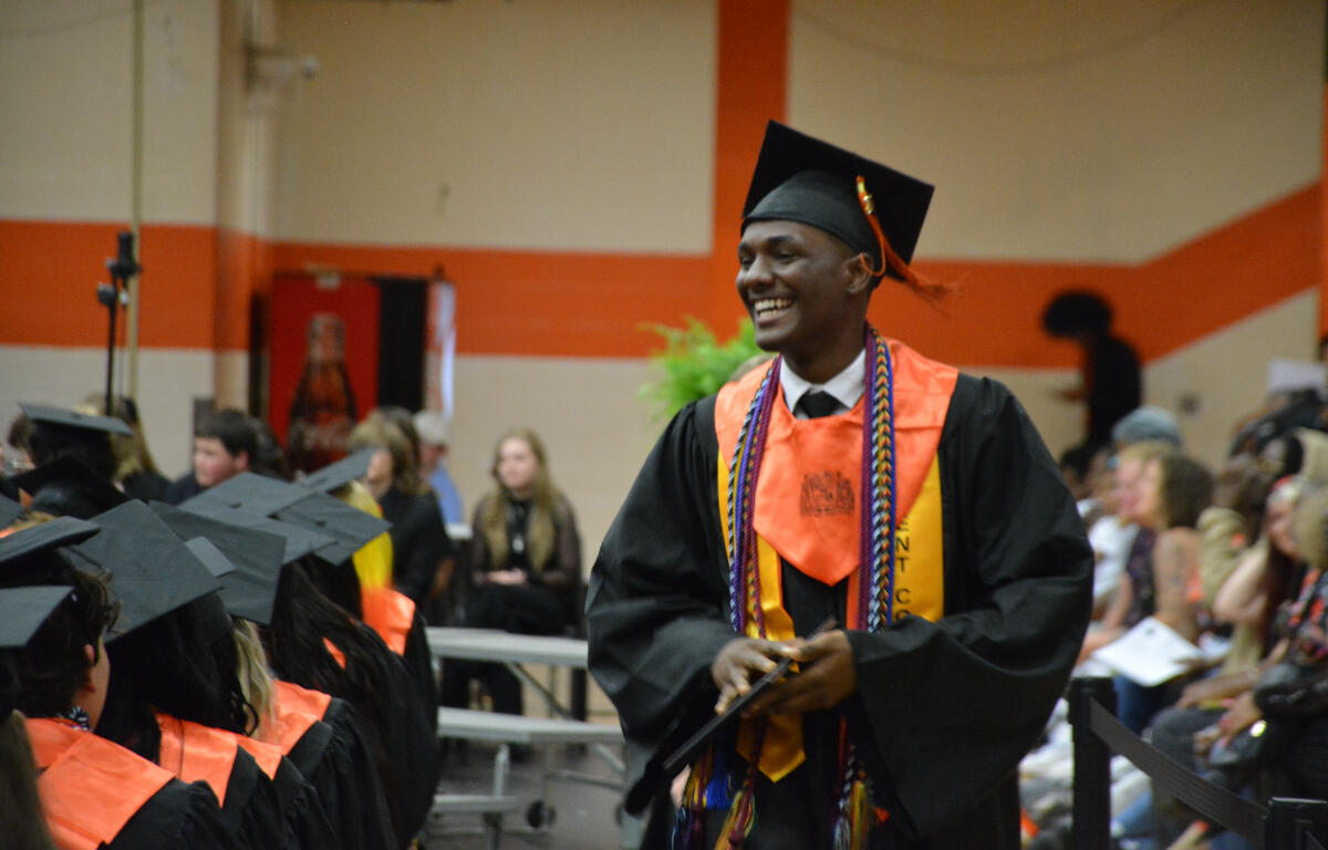Male graduate in black gown and orange stole, smiling as he receives a diploma at a graduation ceremony.