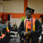 Male graduate in black gown and orange stole, smiling as he receives a diploma at a graduation ceremony.