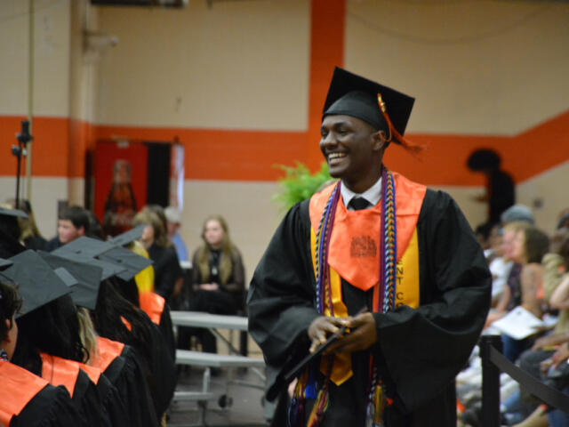 Male graduate in black gown and orange stole, smiling as he receives a diploma at a graduation ceremony.