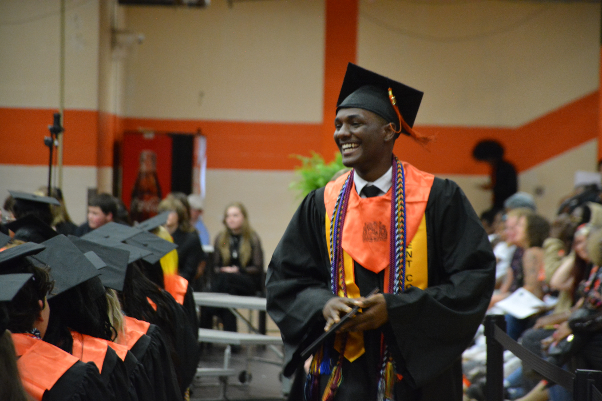Male graduate in black gown and orange stole, smiling as he receives a diploma at a graduation ceremony.