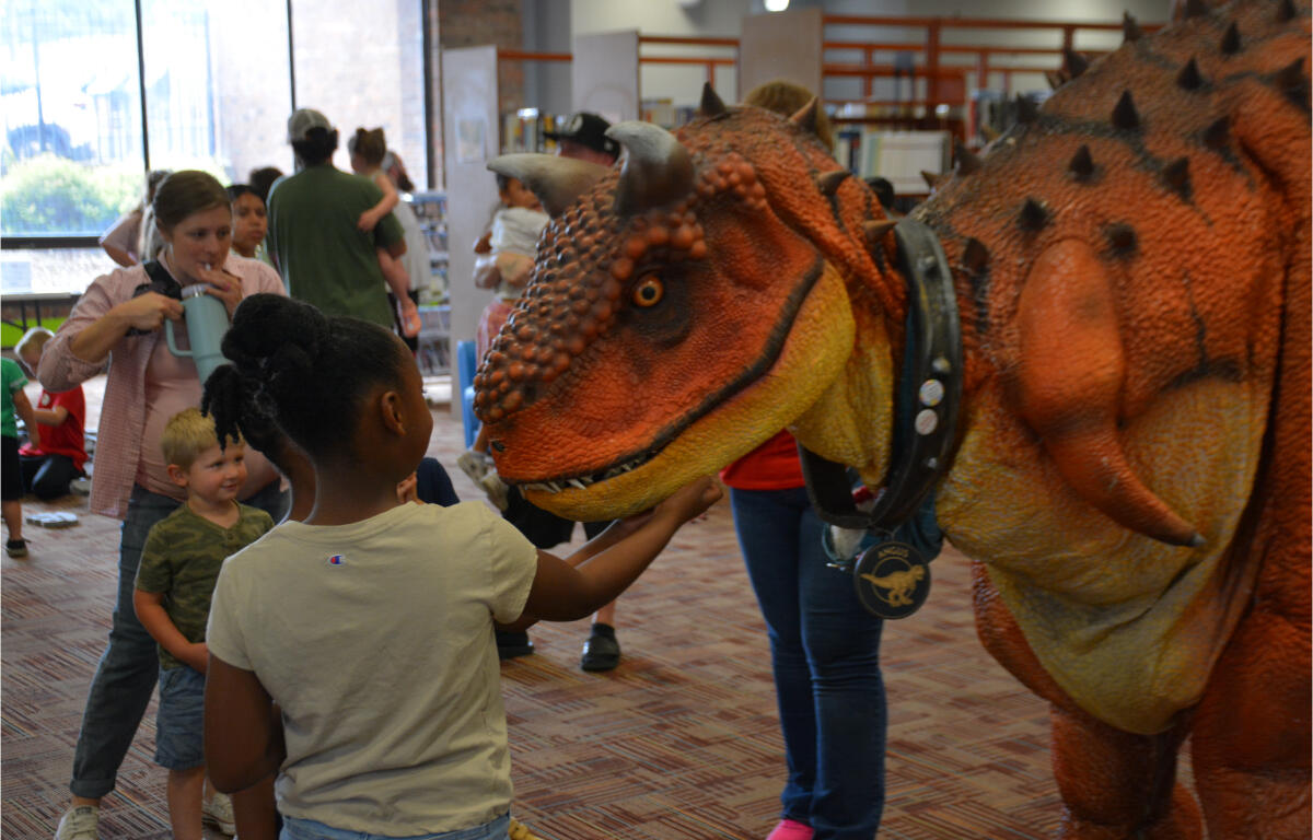 A young girl touches a large orange dinosaur puppet at a crowded library event.