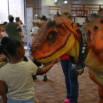 A young girl touches a large orange dinosaur puppet at a crowded library event.