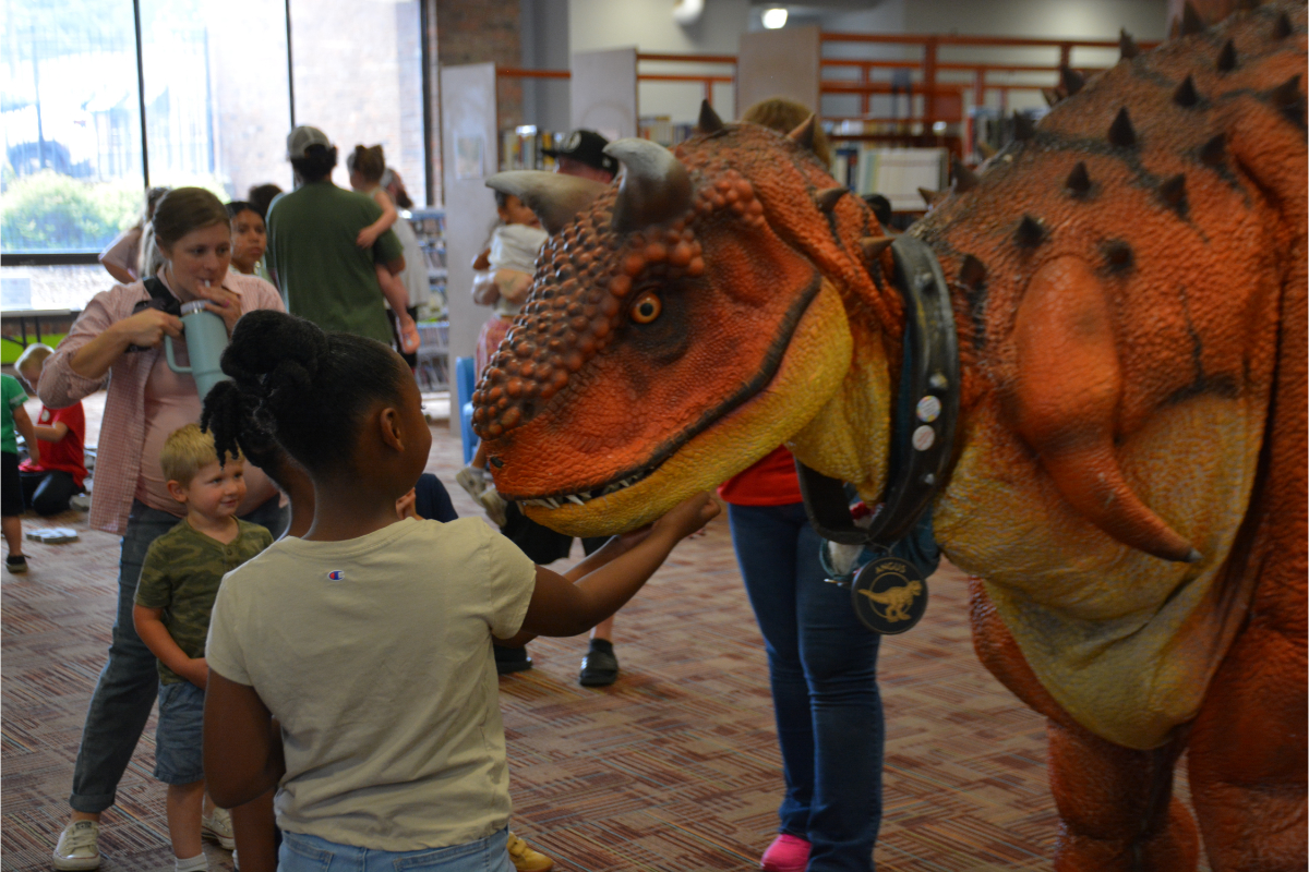 A young girl touches a large orange dinosaur puppet at a crowded library event.