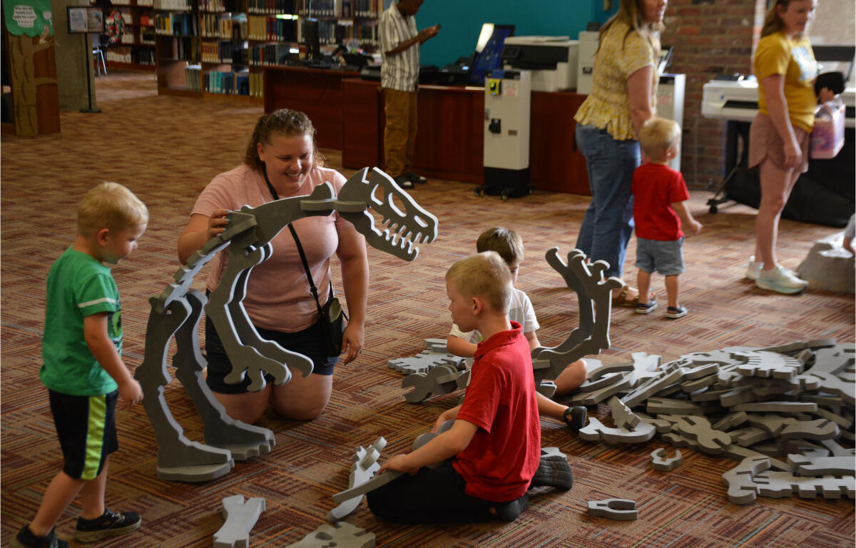 Children and adult kneel on a library floor assembling a large gray foam dinosaur skeleton puzzle.