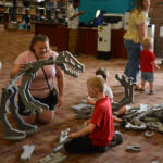 Children and adult kneel on a library floor assembling a large gray foam dinosaur skeleton puzzle.