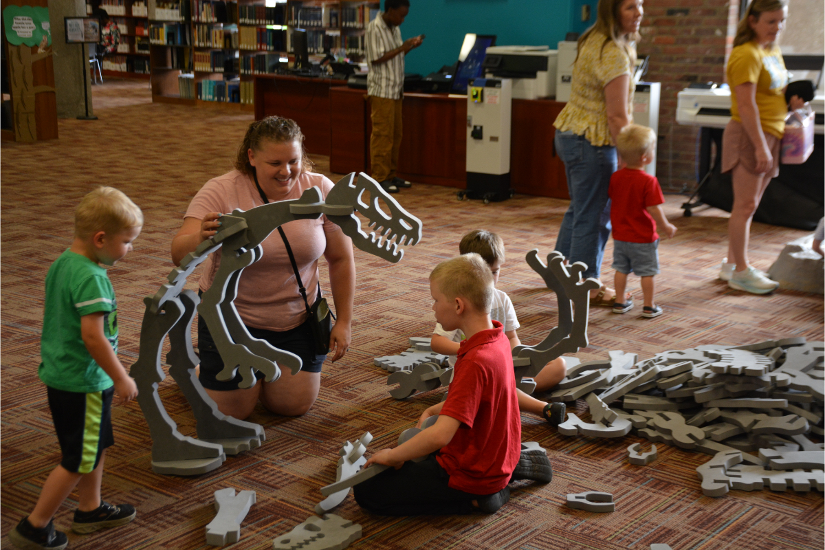 Children and adult kneel on a library floor assembling a large gray foam dinosaur skeleton puzzle.