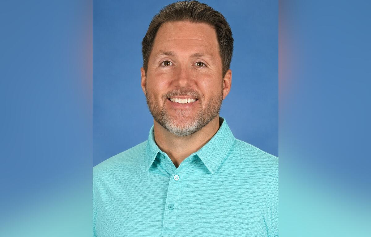 Smiling man with short brown hair and stubble wearing a turquoise polo against a blue backdrop.