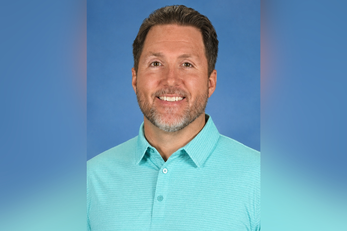 Smiling man with short brown hair and stubble wearing a turquoise polo against a blue backdrop.
