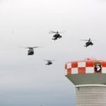 Six helicopters fly in a loose formation above a red-and-white checkered air tower with an Airborne insignia patch at the top.