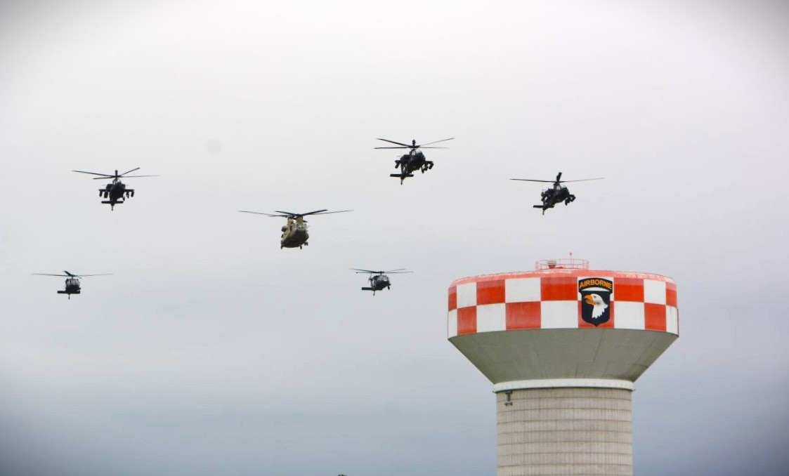 Six helicopters fly in a loose formation above a red-and-white checkered air tower with an Airborne insignia patch at the top.
