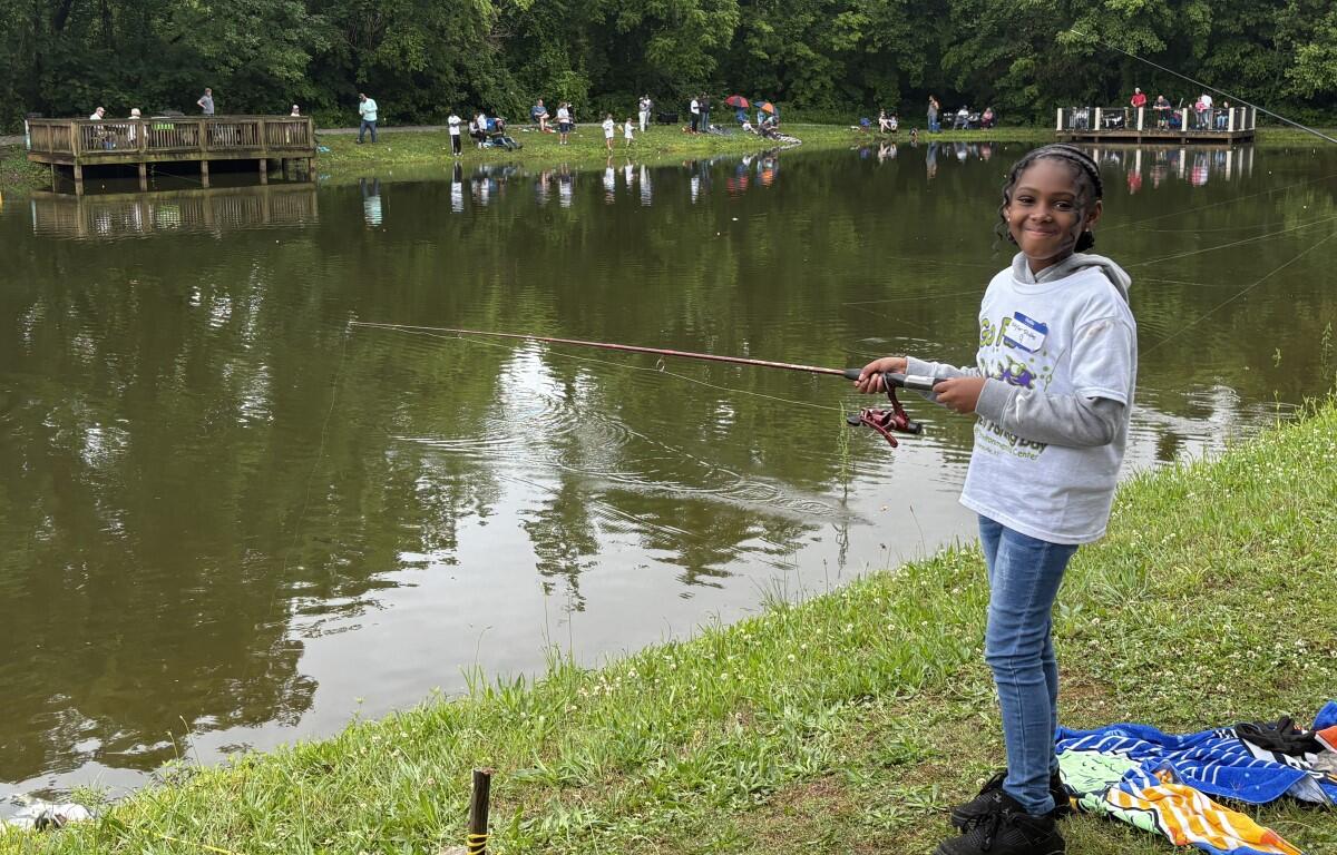 Young girl fishing by a pond, smiling, holding a pink fishing rod with spectators on the far bank.
