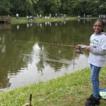 Young girl fishing by a pond, smiling, holding a pink fishing rod with spectators on the far bank.
