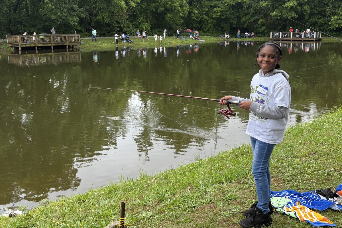 Young girl fishing by a pond, smiling, holding a pink fishing rod with spectators on the far bank.