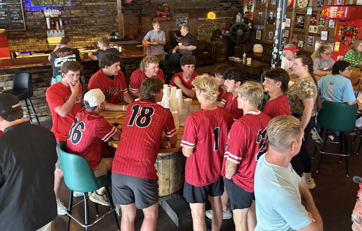 Group of baseball players in red jerseys sitting around a wooden barrel table in a bar, chatting and watching each other.
