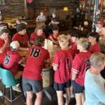 Group of baseball players in red jerseys sitting around a wooden barrel table in a bar, chatting and watching each other.