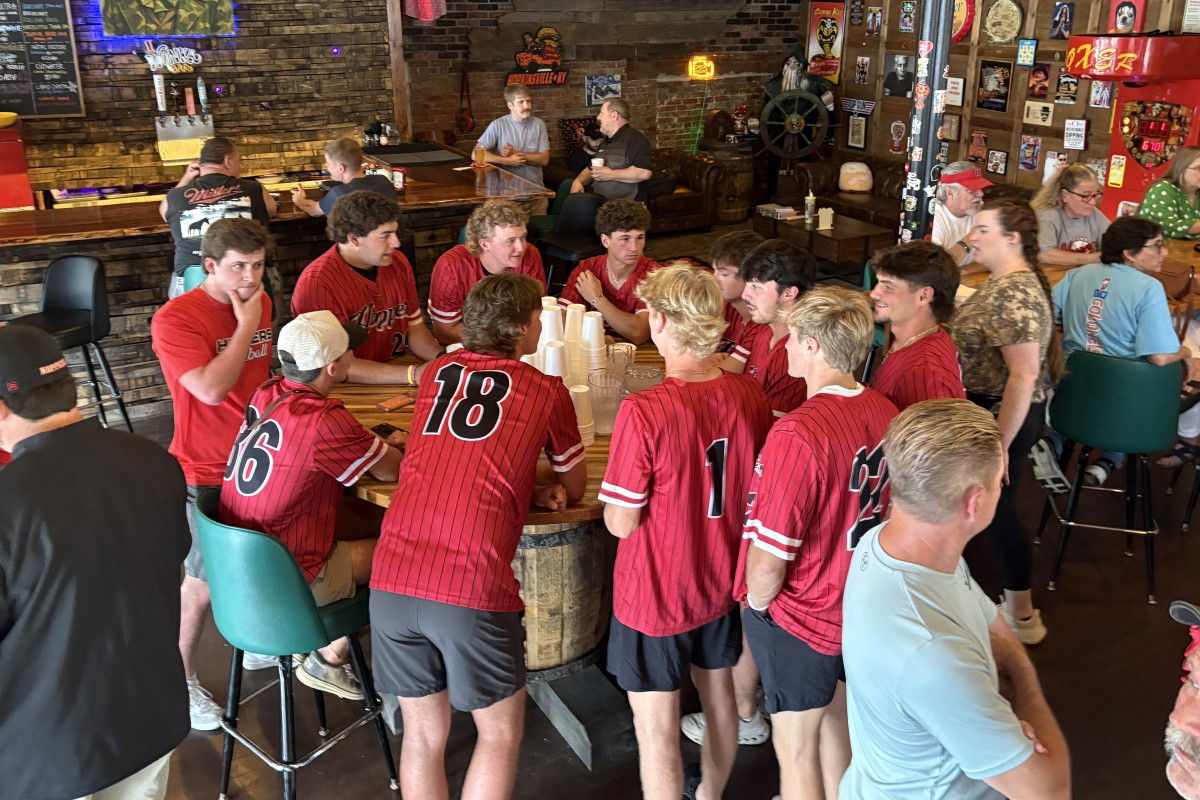 Group of baseball players in red jerseys sitting around a wooden barrel table in a bar, chatting and watching each other.