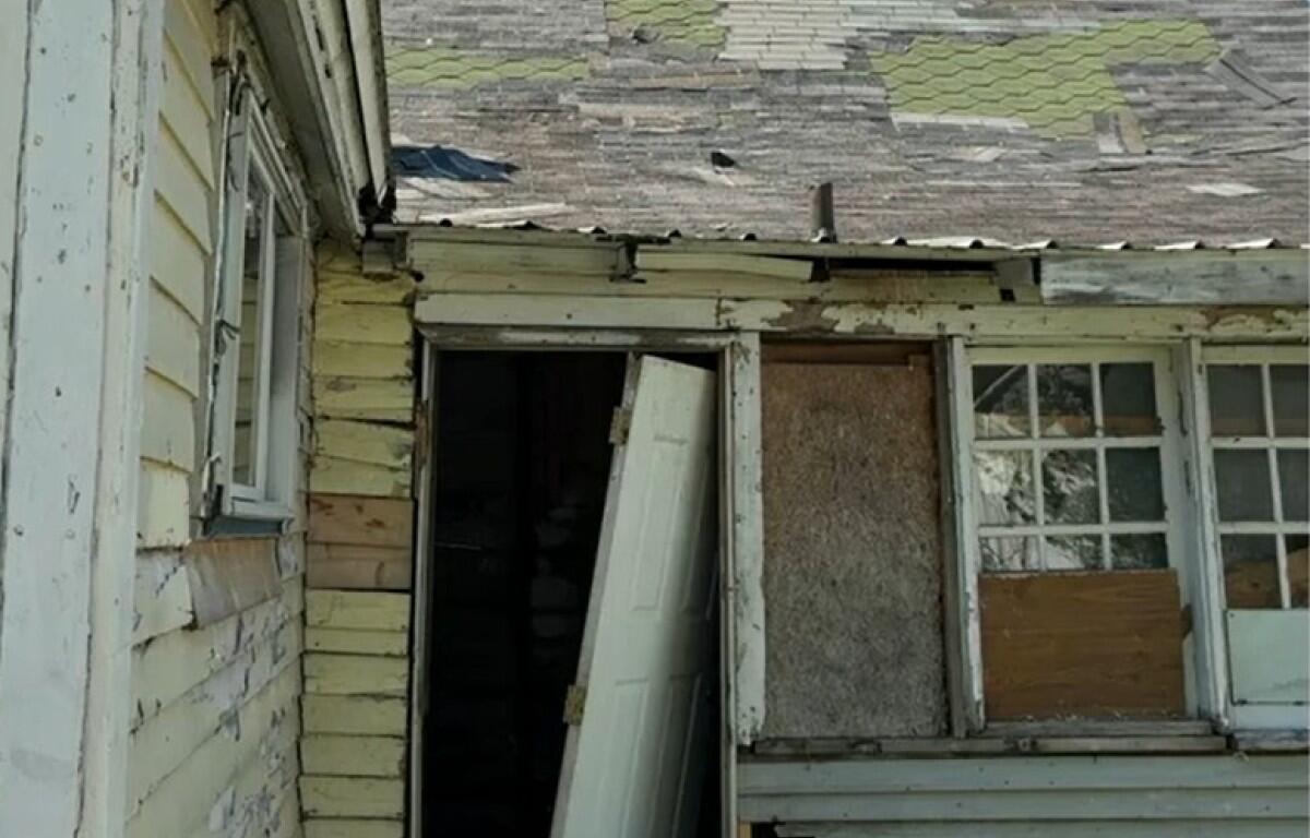Dilapidated house exterior with peeling paint, an ajar door, and a boarded-up window.