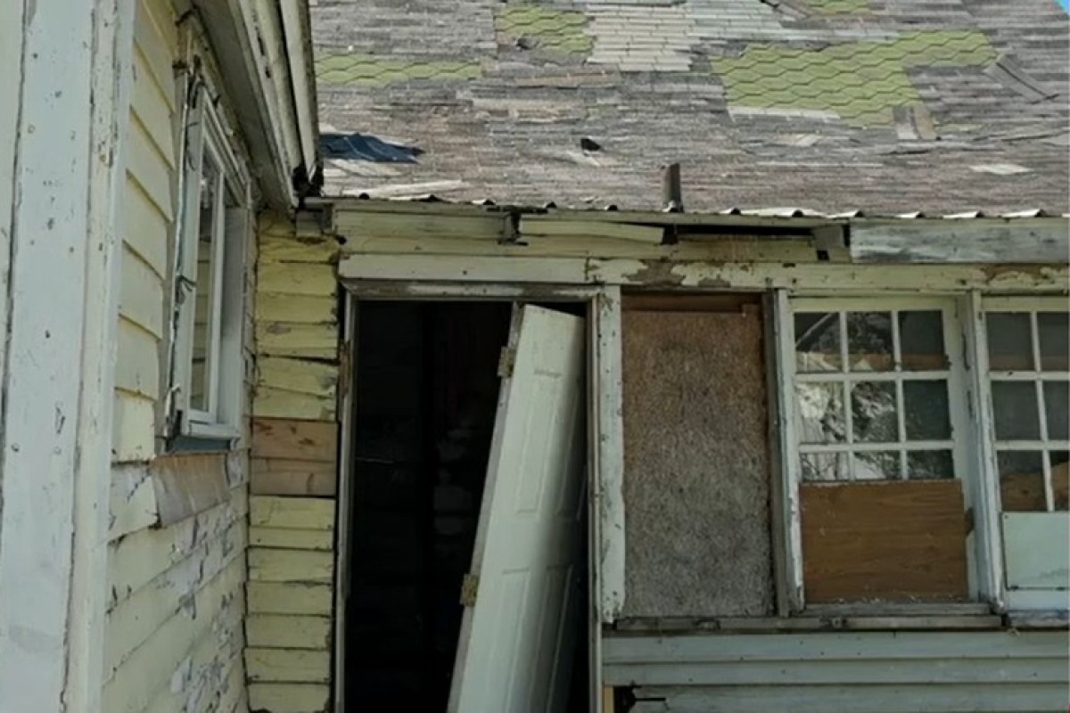 Dilapidated house exterior with peeling paint, an ajar door, and a boarded-up window.