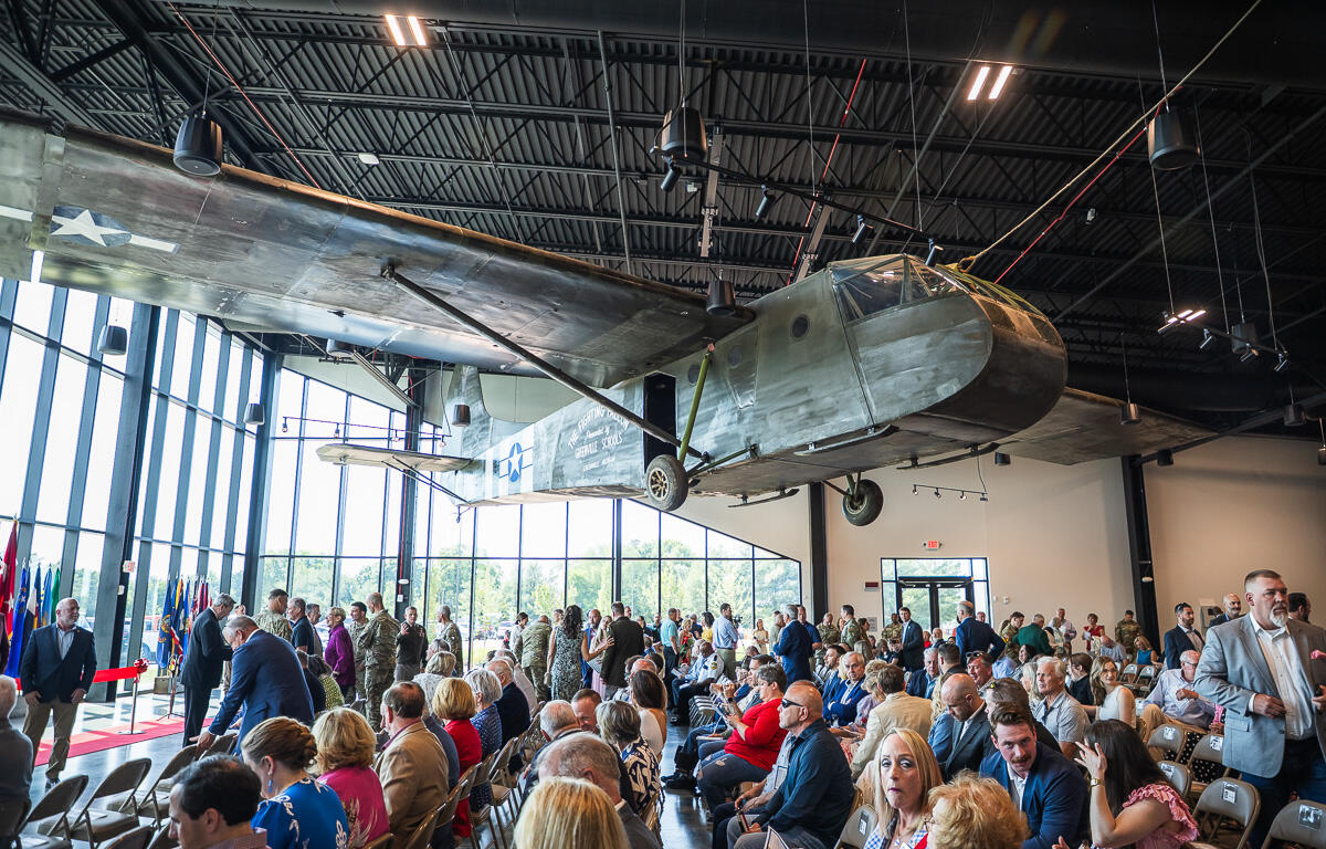 Large vintage military airplane suspended from the ceiling in a hangar, as attendees sit and stand for a ceremony nearby.