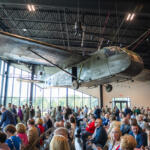 Large vintage military airplane suspended from the ceiling in a hangar, as attendees sit and stand for a ceremony nearby.