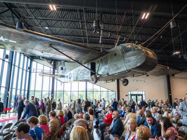 Large vintage military airplane suspended from the ceiling in a hangar, as attendees sit and stand for a ceremony nearby.