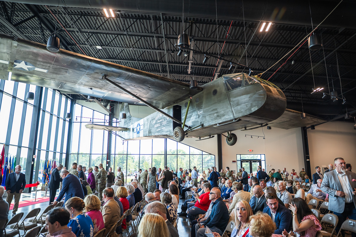 Large vintage military airplane suspended from the ceiling in a hangar, as attendees sit and stand for a ceremony nearby.