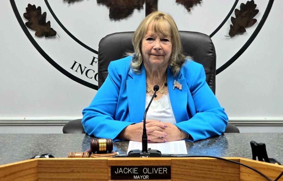 Public official in a bright blue blazer sits at a council desk with a microphone and gavel nearby.