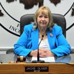 Public official in a bright blue blazer sits at a council desk with a microphone and gavel nearby.