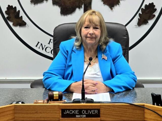 Public official in a bright blue blazer sits at a council desk with a microphone and gavel nearby.