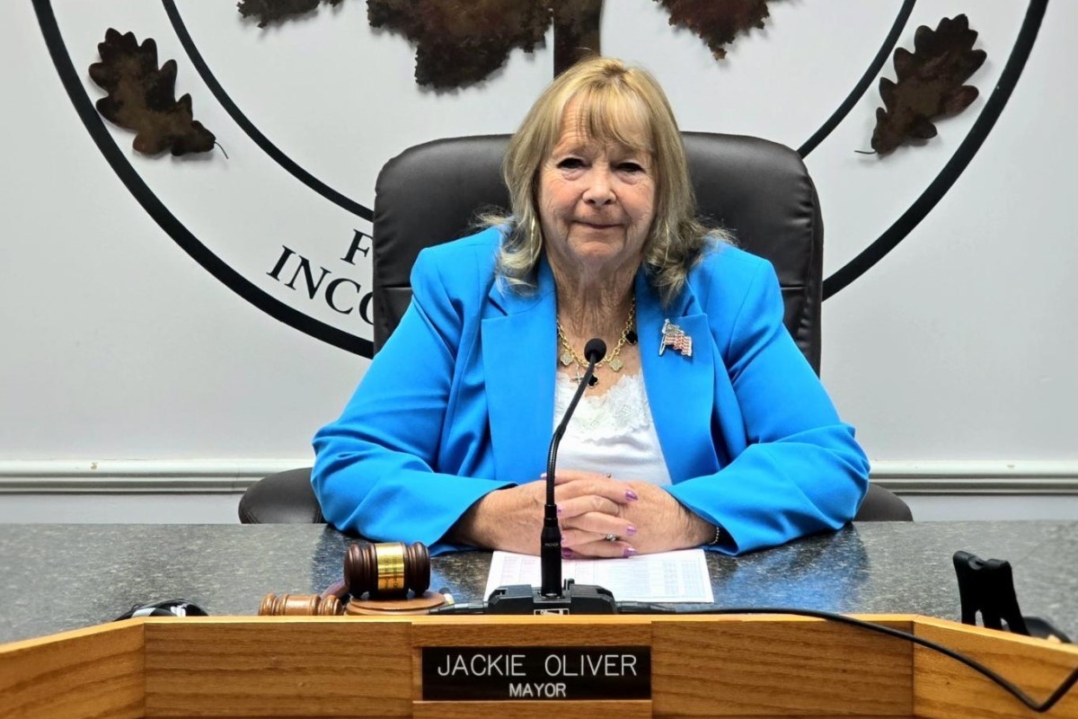 Public official in a bright blue blazer sits at a council desk with a microphone and gavel nearby.