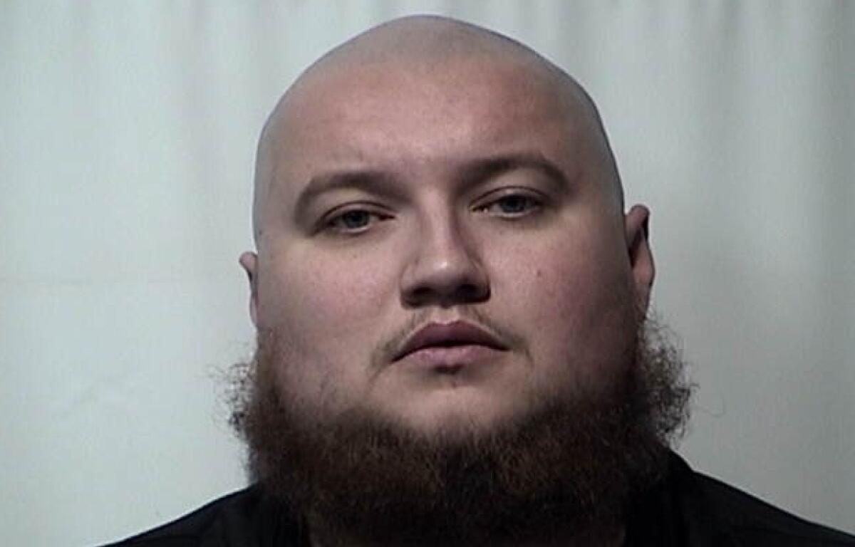 Mugshot of a bald man with a thick beard facing the camera against a plain light background.