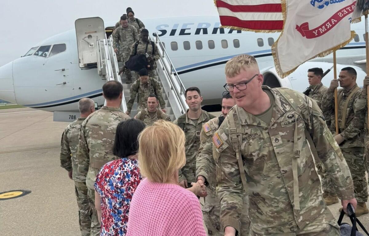 U.S. Army soldiers in camouflage greet civilians on the tarmac after exiting a military aircraft, with a flag to the side.