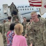U.S. Army soldiers in camouflage greet civilians on the tarmac after exiting a military aircraft, with a flag to the side.