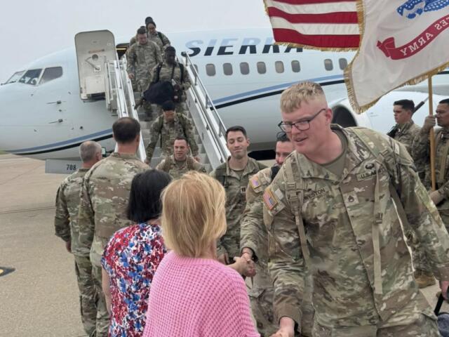 U.S. Army soldiers in camouflage greet civilians on the tarmac after exiting a military aircraft, with a flag to the side.