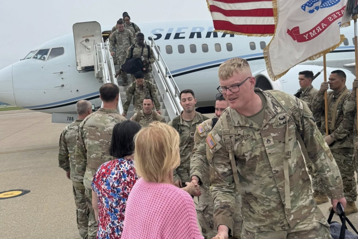 U.S. Army soldiers in camouflage greet civilians on the tarmac after exiting a military aircraft, with a flag to the side.