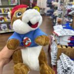 Person holds a plush mascot wearing a blue shirt with a circular cartoon dog badge in a toy store background shelves and clothes