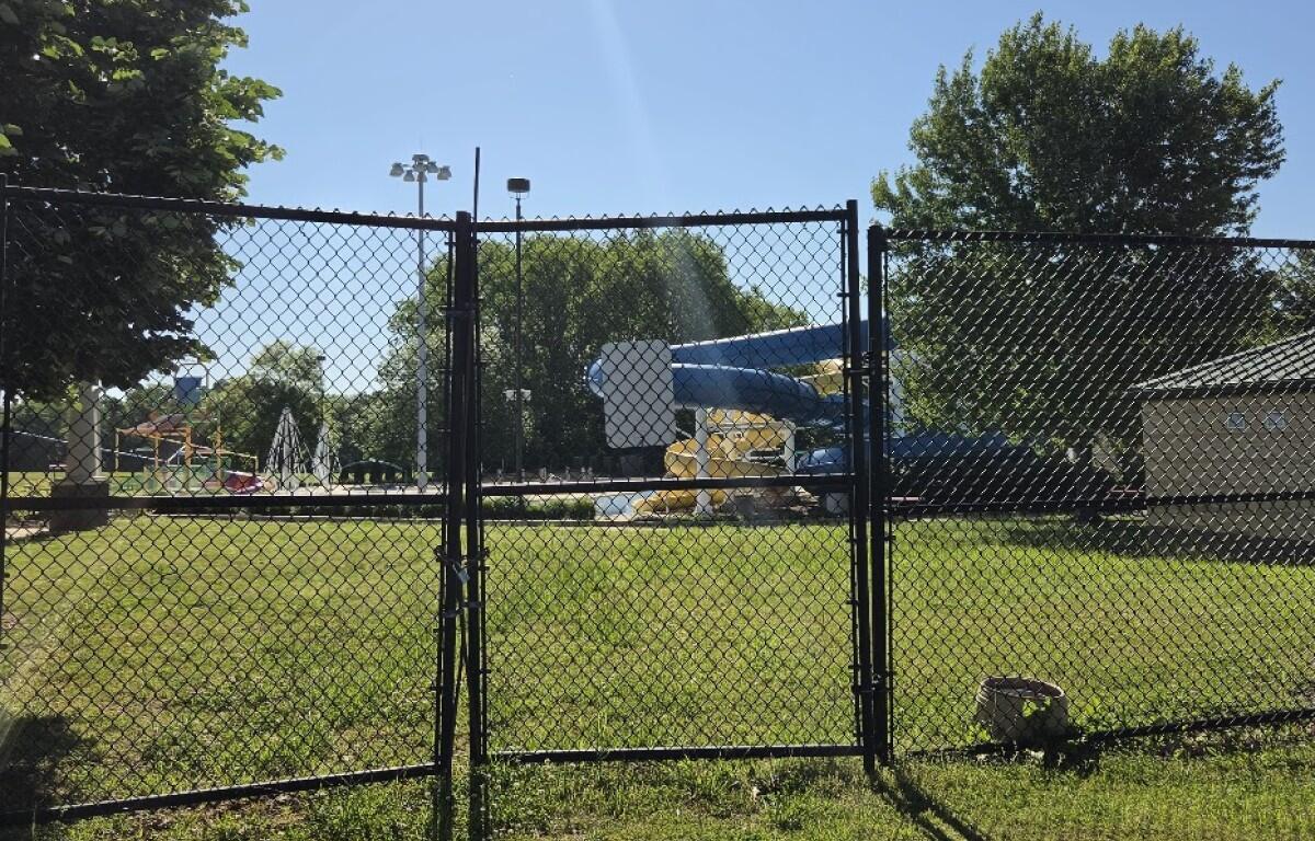 Gate opens to a park playground with a blue slide and yellow supports behind a chain-link fence on a sunny day.